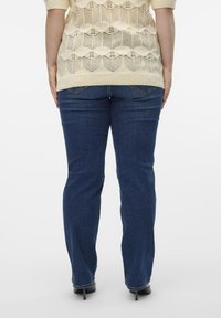 Lichtbeige gebreide trui met een open weefpatroon, gedragen over donkerblauwe rechte jeans, gecombineerd met zwarte hoge hakken.