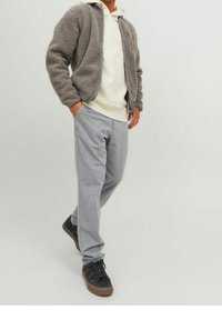 Giacca grigia e pelosa con zip sopra un hoodie bianco, abbinata a pantaloni grigio chiaro a righe sottili e sneakers nere con suola in gomma.