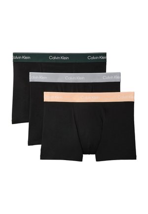 Atletyczny czarnoskóry mężczyzna stoi boso na szarym tle, nosząc czarne bokserki Calvin Klein, ręce za plecami.