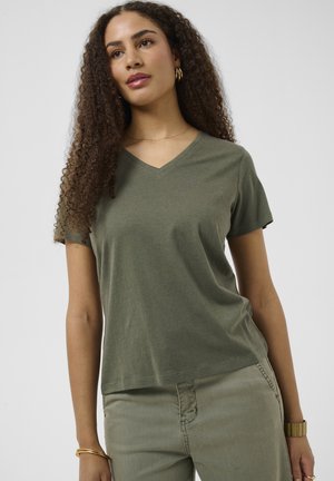 Jeune femme aux longs cheveux bouclés portant un t-shirt vert à col en V et un pantalon vert clair, accessoirisée avec des bijoux en or et une montre.