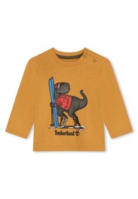 Langdurige mouw mosterdkleurig shirt met een cartoon dinosaur met zonnebril, ski's en laarzen. Bevat Timberland-logo en grafische details.