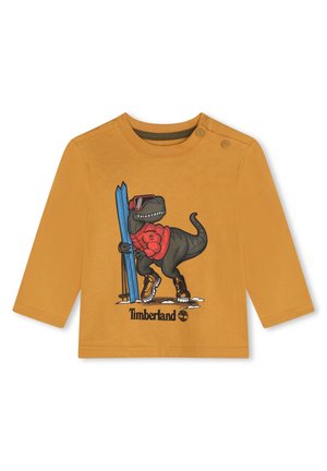 Camicia a maniche lunghe color senape con un dinosauro cartone animato che indossa occhiali da sole, sci e stivali. Include il logo Timberland e dettagli grafici.