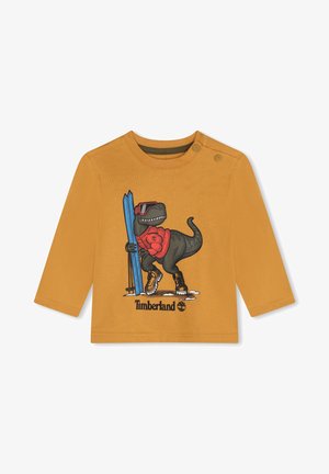 Camicia a maniche lunghe color senape con un dinosauro cartone animato che indossa occhiali da sole, sci e stivali. Include il logo Timberland e dettagli grafici.