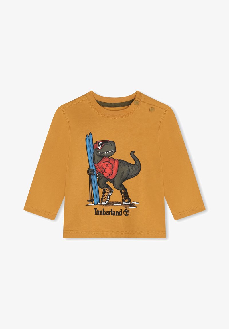 Chemise à manches longues couleur moutarde avec un dinosaure de dessin animé portant des lunettes de soleil, des skis et des bottes. Comprend le logo Timberland et des détails graphiques.