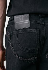 Jean en denim noir avec des détails déchirés, présentant une étiquette en cuir noir texturé sur la ceinture. Gros plan sur la poche arrière.