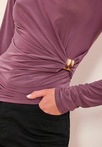 Langærmet top i mauve stof med rynket tekstur ved taljen og en guldfarvet spænde som detalje. Sorte bukser med en synlig sidelomme.