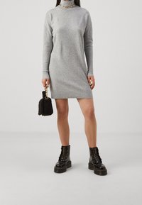 Femme portant une robe tricotée grise à manches longues, des bottes noires à lacets épaisses, un collier en chaîne dorée, tenant un sac à main en fausse fourrure noire avec une anse en chaîne dorée.