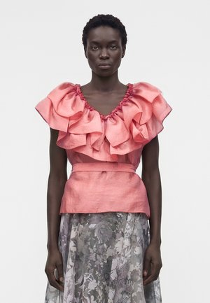 Kobieta ubrana w błyszczącą różową bluzkę z falbanami, bufkami i dopasowaną talią, zestawioną z przezroczystą spódnicą w kwiatowy wzór.