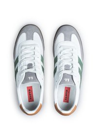 Zapatillas blancas con rayas verdes, detalles en gamuza gris y una suela de goma marrón. Presentan cordones redondos y una etiqueta prominente en el interior.