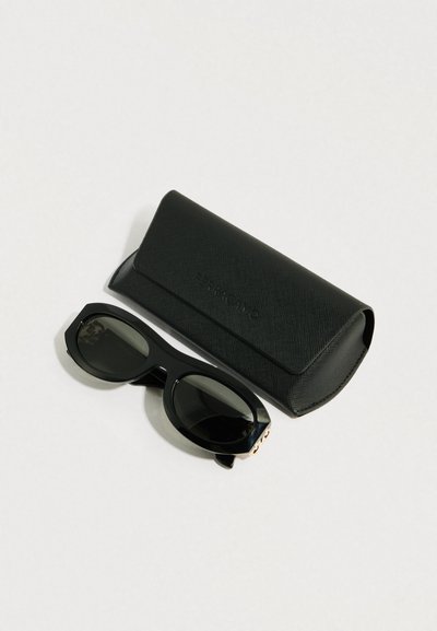 Μαύρα γυαλιά ηλίου με χοντρό, γωνιώδες πλαίσιο και σκουρόχρωμους φακούς, τοποθετημένα δίπλα σε μαύρη θήκη προστασίας με υφή και κλείσιμο με καπάκι.