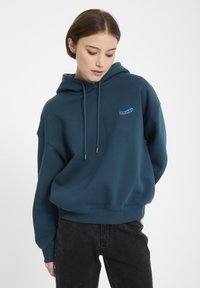 Turkusowa bluza z kapturem z miękkiego materiału o luźnym kroju, z logo z przodu i regulowanymi sznurkami. Noszona z czarnymi dżinsami.