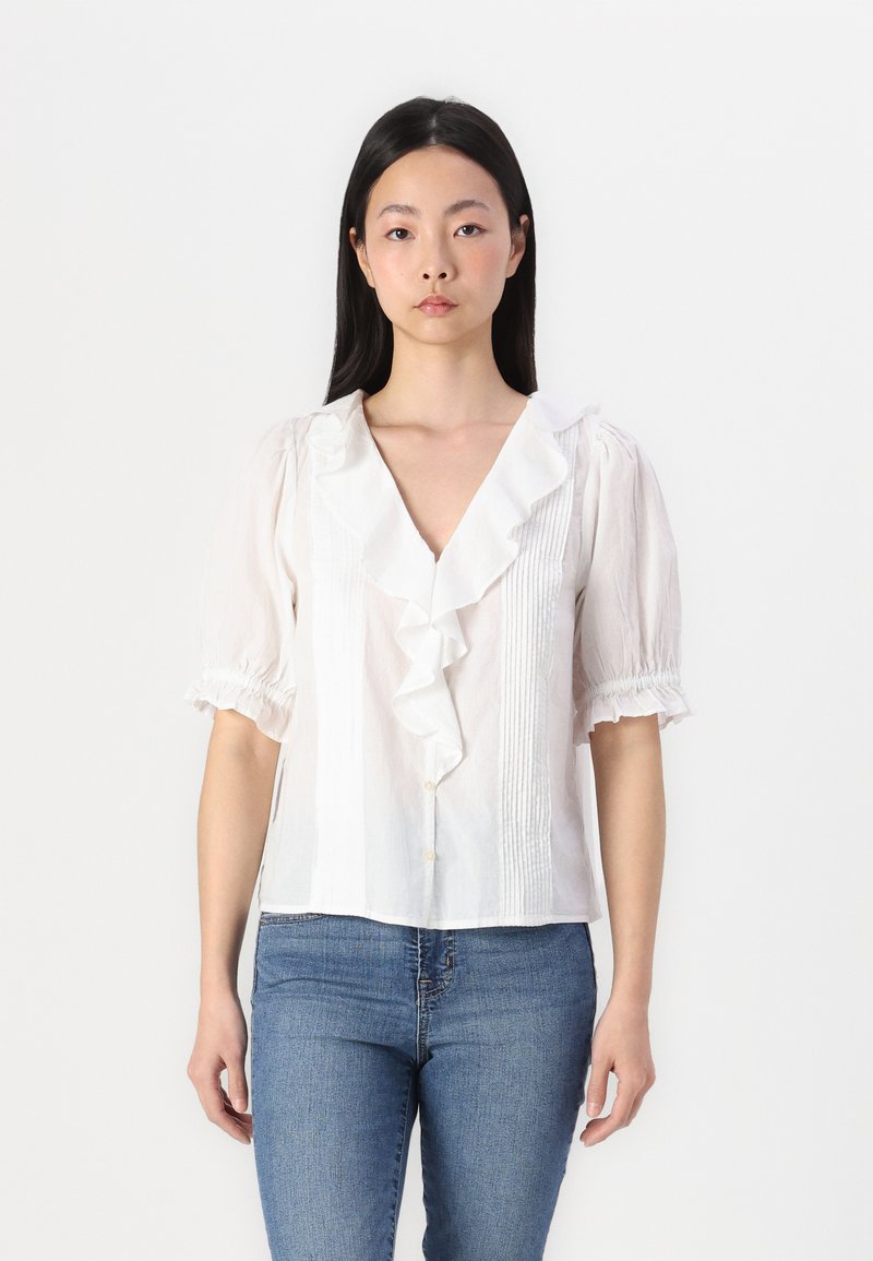Biele blouse s volánovým golierom, krátkymi puff rukávmi a plisovanými detailmi. Obsahuje zapínanie na gombíky a voľný strih. Kombinovaná s modrými džínami.
