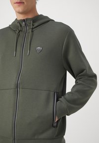 Felpa con zip di color verde oliva, realizzata in tessuto morbido, con un piccolo logo, due tasche laterali con zip nera e cappuccio regolabile con cordino.