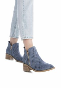 Stivali alla caviglia blu perforati con cerniere laterali, punta rotonda e tacco marrone accatastato, abbinati a jeans di denim chiaro.