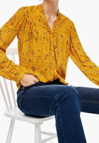 Personne assise sur une chaise blanche portant un chemisier jaune moutarde à motifs, boutonné, et un jean bleu foncé, avec une main dans la poche.