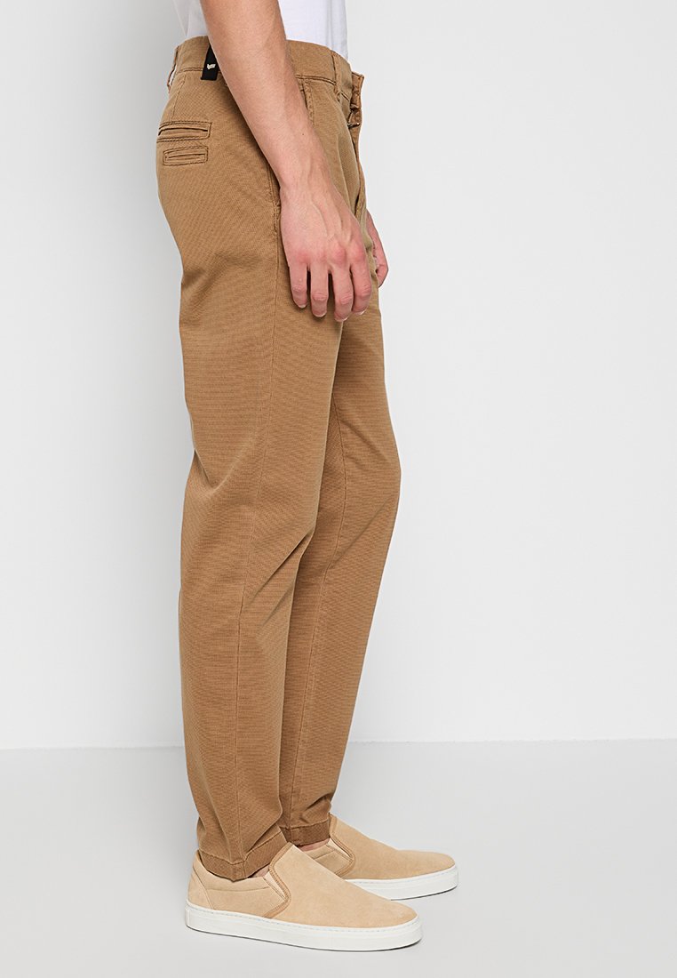 Mann in schmalen braunen Hosen und beigen Slippern, der vor einem einfarbigen hellgrauen Hintergrund steht.