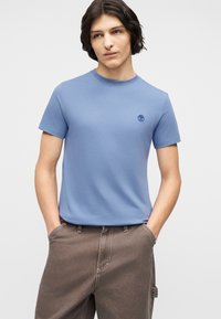 Νέος άνδρας με μακριά μέχρι τους ώμους σκούρα μαλλιά, φορώντας ανοιχτό μπλε κοντομάνικο πουκάμισο και καφέ παντελόνι, με τα χέρια στις τσέπες.