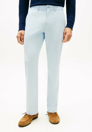 Mężczyzna ubrany w jasnoniebieskie spodnie, granatową bluzę z długim rękawem oraz brązowe zamszowe buty sznurowane, stojący z rękami swobodnie opuszczonymi wzdłuż ciała.