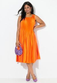 Vestido naranja plisado con escote redondo, combinado con un bolso morado de lentejuelas y tacones lavanda, destacando por su textura suave y colores vibrantes.