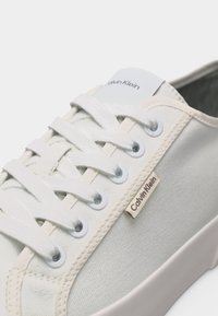 Λευκό πάνινο αθλητικό παπούτσι με λευκά κορδόνια, μεταλλικά κρικάκια και ετικέτα με το λογότυπο Calvin Klein στο πλάι κοντά στα κορδόνια.