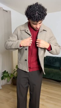 Beige takki, jossa on kaulus, edessä napit ja sileä pinta. Alla punainen neulepaita. Tummat housut ja sisätilat.