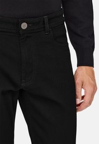 Černé džíny z denimu s klasickým designem, vyznačující se kovovým knoflíkem, viditelným švem a standardním pasem. Ruka spočívá na boku.