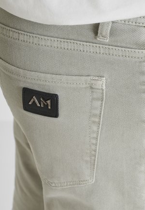 Close-up van de achterzak van lichtgrijze broek met een zwarte patch met metalen "AM"-logo op gestikte stof.