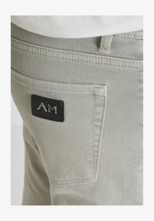 Close-up of light gray pants back pocket with black patch featuring metallic "AM" logo on stitched fabric.