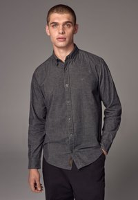 Camicia button-up grigia scura in tessuto testurizzato, con design con colletto, maniche lunghe e un logo discreto vicino al petto. Pantaloni neri.