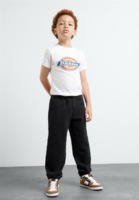 Balta trumpa rankovėmis marškinėliai su mėlynu ir oranžiniu "Dickies" logotipu, derinami su juodomis sportinėmis kelnėmis ir rudos bei baltos spalvos sportbačiais.