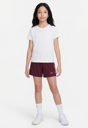Jeune fille debout, portant un t-shirt blanc, un short de sport bordeaux avec des accents rouges, des chaussettes blanches montantes et des baskets blanches, sur un fond uni.