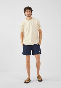 T-shirt beige in cotone, pantaloni da bagno blu navy e sandali marroni. I pantaloni presentano un logo, mentre la maglietta ha maniche corte e una vestibilità comoda.