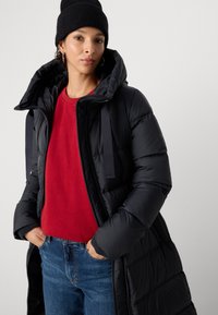 Zwarte pufferjas met oversized capuchon, ritsluiting en lintaccenten, gedragen over een rode trui en gecombineerd met blauwe jeans.