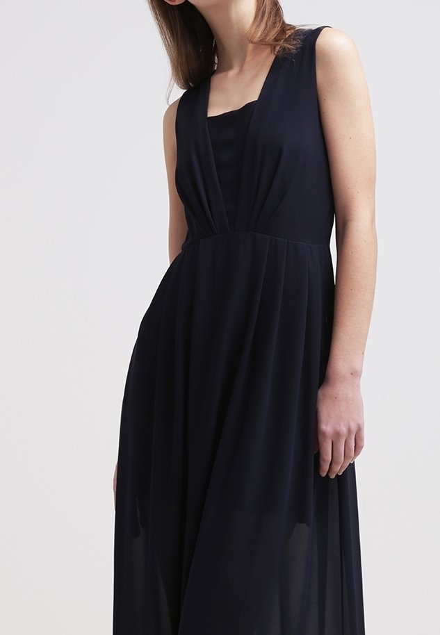 Femme portant une robe noire sans manches avec un profond décolleté en V et une taille froncée sur un fond clair uni.