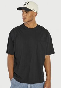 Camiseta negra de manga corta con cuello redondo y un pequeño bolsillo en el pecho, combinada con una gorra de béisbol beige y negra que presenta un logo.