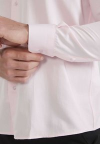 Chemise boutonnière rose pâle avec une texture lisse, présentant un unique bouton de poignet et une coupe décontractée, partiellement rentré dans un pantalon noir.