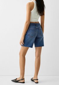 Pantalones cortos de mezclilla en un tono azul medio con dobladillo deshilachado, combinados con un top blanco sin mangas y sandalias planas negras, con un diseño sencillo.