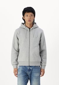 Grå zip-up hoodie med en frontlomme og justerbar snoretrækshætte, lavet af blødt stof. Båret med lyseblå jeans.