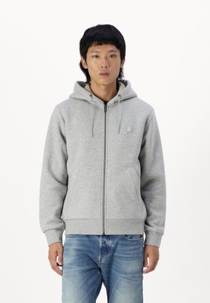 Grå zip-up hoodie med en frontlomme og justerbar snoretrækshætte, lavet af blødt stof. Båret med lyseblå jeans.