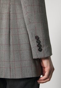 Blazer de cuadros gris con acentos rojos; presenta cuatro botones negros en el puño; tejido texturizado con patrones de cuadros distintivos.