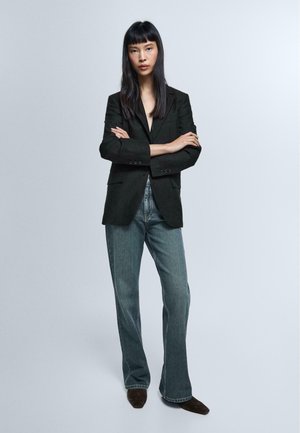 Frau mit langen schwarzen Haaren, die einen schwarzen Blazer, blaue Jeans und braune spitze Schuhe trägt und mit verschränkten Armen vor einem schlichten Hintergrund steht.