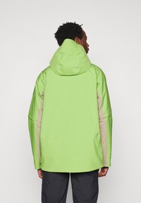 Chaqueta impermeable verde con capucha, con acentos beige en los laterales y las mangas, textura suave y ajuste holgado.