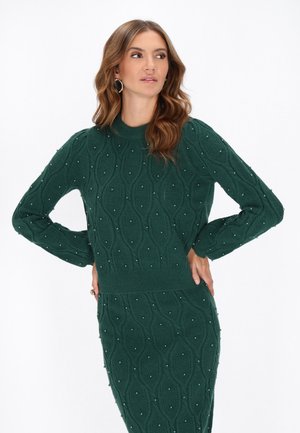 Donna che indossa un completo maglione e gonna di colore verde scuro con piccoli dettagli di perline, in piedi con le mani sui fianchi e che guarda di lato.