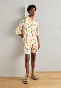 Camisa de manga corta y de corte holgado en beige, con patrones tropicales coloridos. Combinada con sandalias beige.