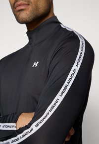 Μαύρο πουλόβερ με φερμουάρ μέχρι τη μέση, κατασκευασμένο από λείο ύφασμα, με λευκή σήμανση "Under Armour" στα μανίκια και ψηλό γιακά.