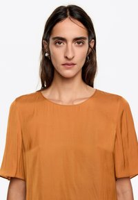 Brązowy top z krótkim rękawem i okrągłym dekoltem. Wykonany z gładkiej tkaniny o lekko luźnym kroju. Bez widocznych wzorów ani zdobień.