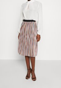 Blouse blanche à manches longues avec un détail en trou de serrure, associée à une jupe plissée midi ornée de bandes verticales colorées. Taille élastique noire.