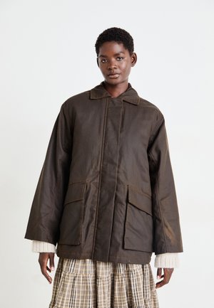 Giacca marrone oversize con colletto strutturato, grandi tasche frontali e cuciture a contrasto; abbinata a una gonna a strati a quadretti.