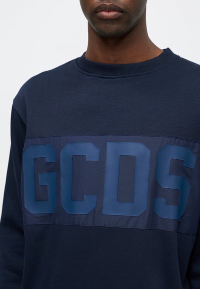 Marinblå sweatshirt med en glansig mörkblå "GCSD"-logotyp över bröstet, med rund hals och en slät textur.