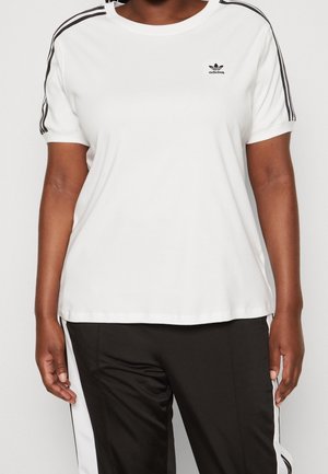 Personne portant un T-shirt blanc Adidas avec des rayures noires sur les manches et un pantalon noir avec des rayures blanches sur les côtés, debout devant un fond uni.